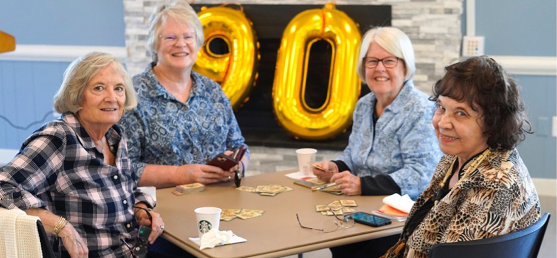 Four older women sit around a table playing cards and smiling. Behind them, large gold balloons form the number 90, suggesting a celebration. Drinks and cards are on the table.