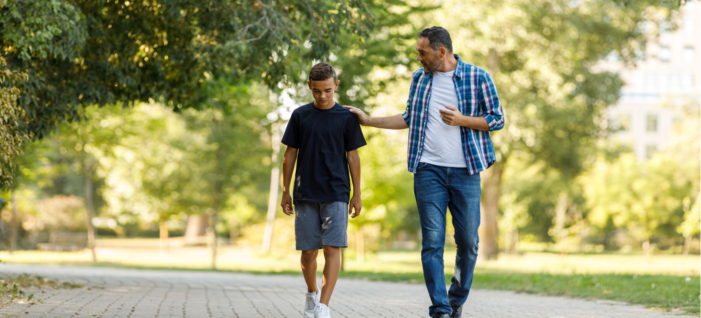 A man and a boy walk together on a park path. The man has his hand on the boys shoulder, and they appear to be having a serious conversation. Trees and greenery surround them on a sunny day.