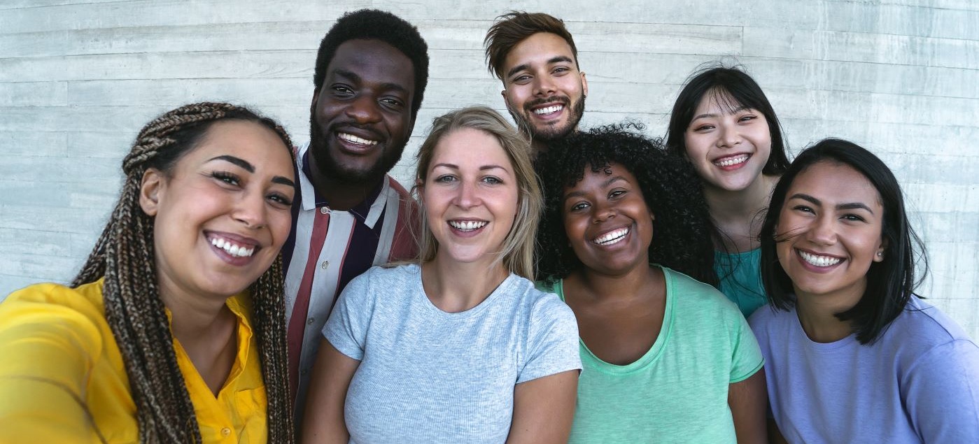 A diverse group of seven young adults stand close together, smiling at the camera, against a plain light gray wall. They appear happy and relaxed, wearing casual, colorful clothing.